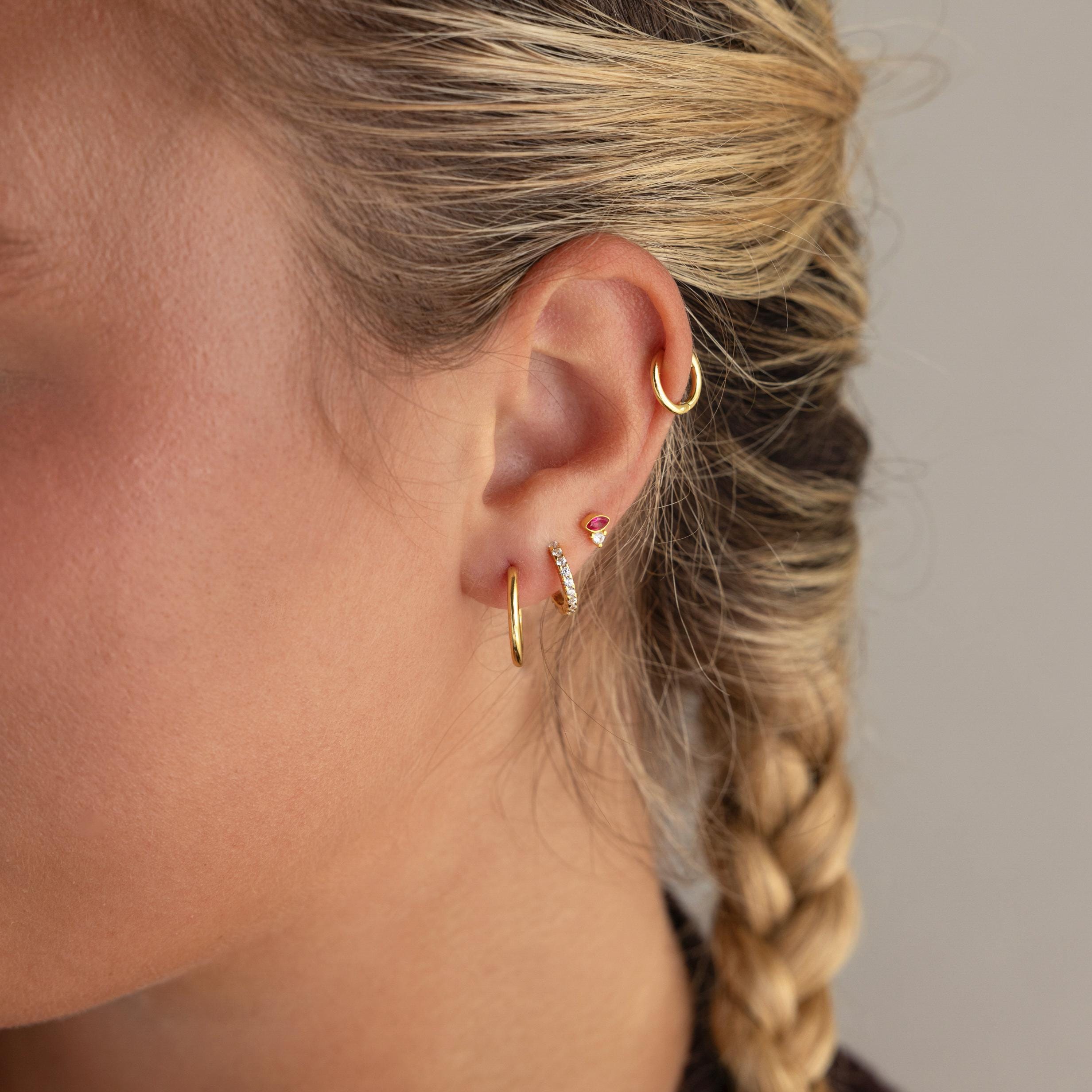 Close-up of a woman's ear adorned with multiple gold earrings, featuring Diamond Marquise Birthstone Studs and a small braid.