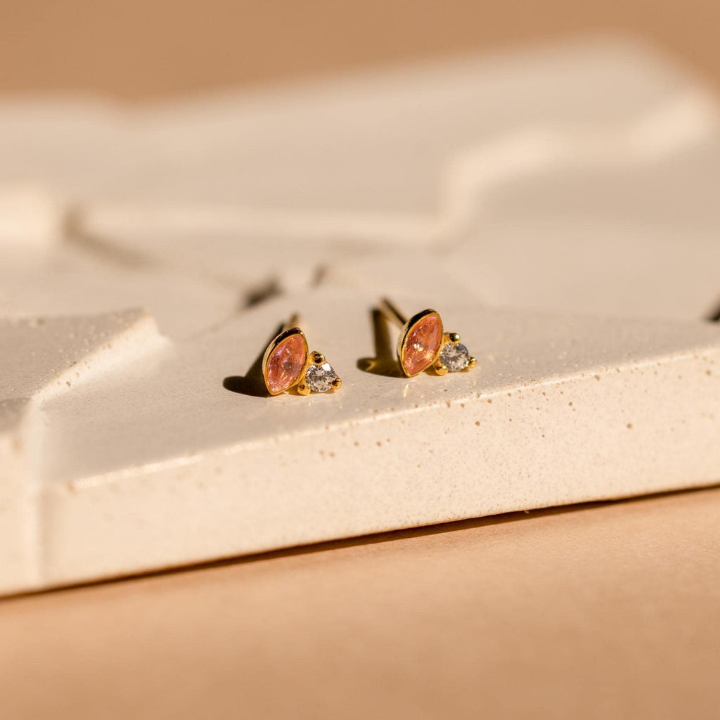 Diamond Marquise Birthstone Studs feature pink oval and clear round stones in a gold setting, adding elegance to any textured cream surface.