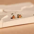 Diamond Marquise Birthstone Studs feature pink oval and clear round stones in a gold setting, adding elegance to any textured cream surface.