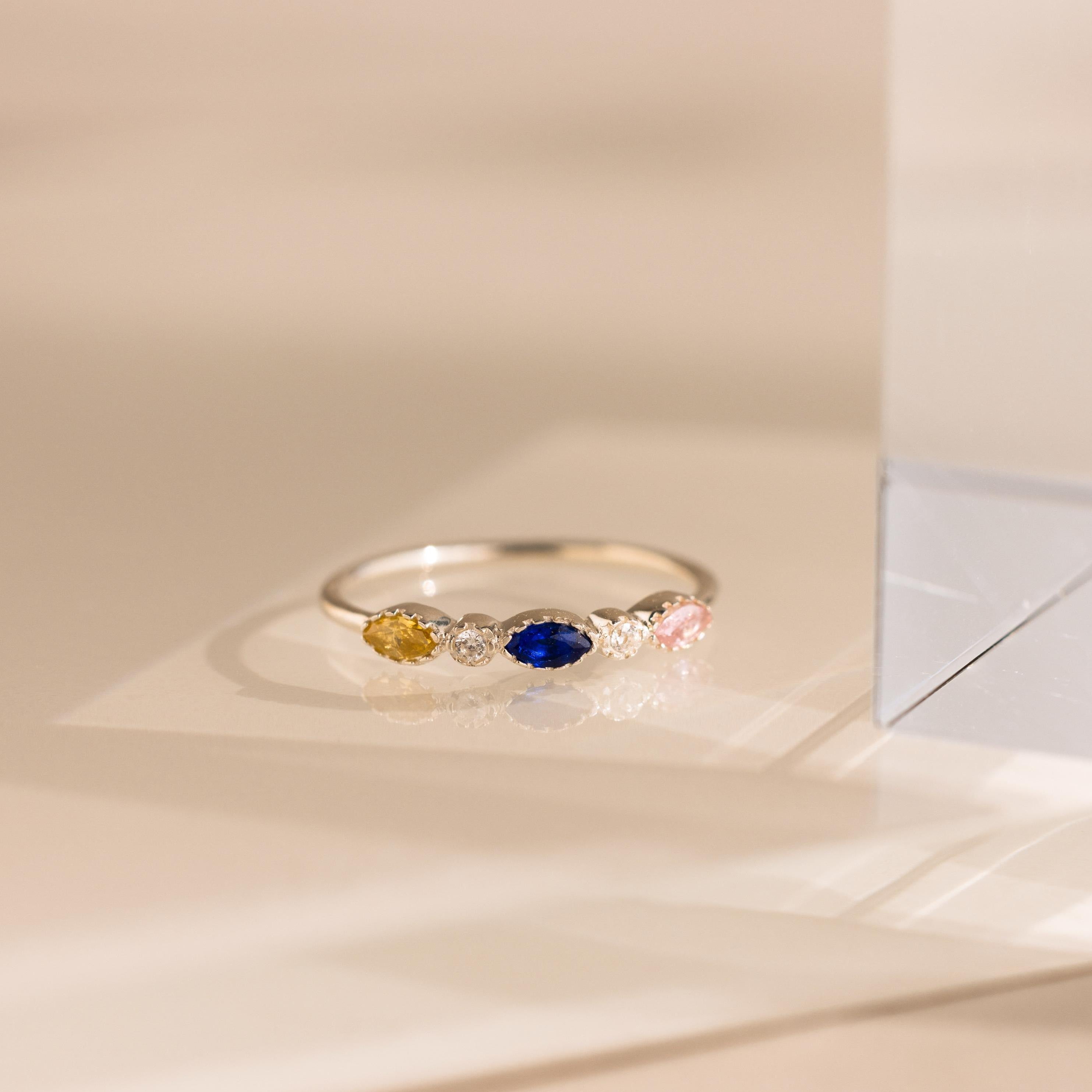 The Cluster Marquise Birthstone Ring in gold features yellow, clear, blue, and pink gemstones that shine beautifully on a reflective surface.