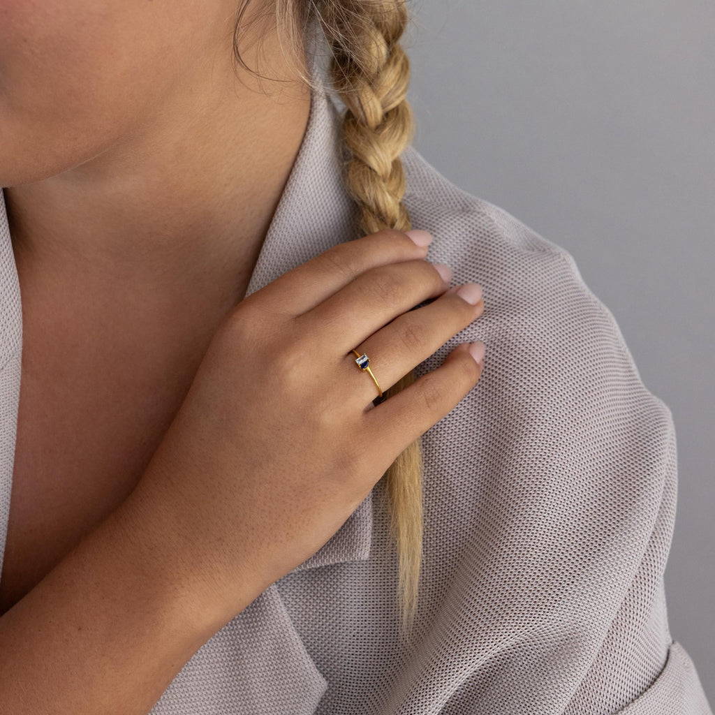 A person with a blonde braid wears the Tiny Toi et Moi Birthstone Ring, featuring a blue stone, with their hand resting on their shoulder.