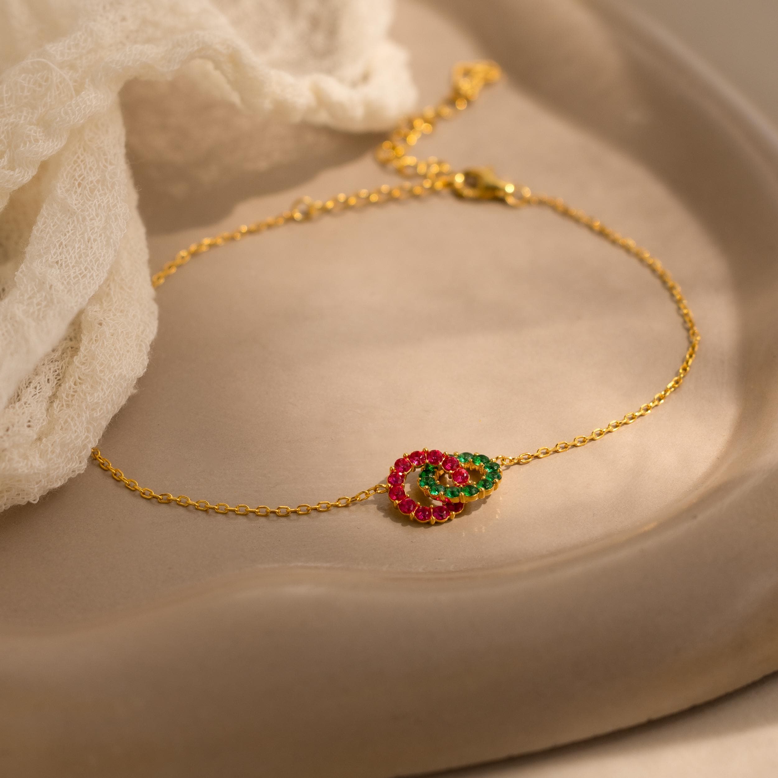 The Interlocking Duo Birthstone Bracelet features customizable red and green gemstones on a delicate gold chain, beautifully displayed on a beige ceramic dish beside lace—an ideal personalized gift.