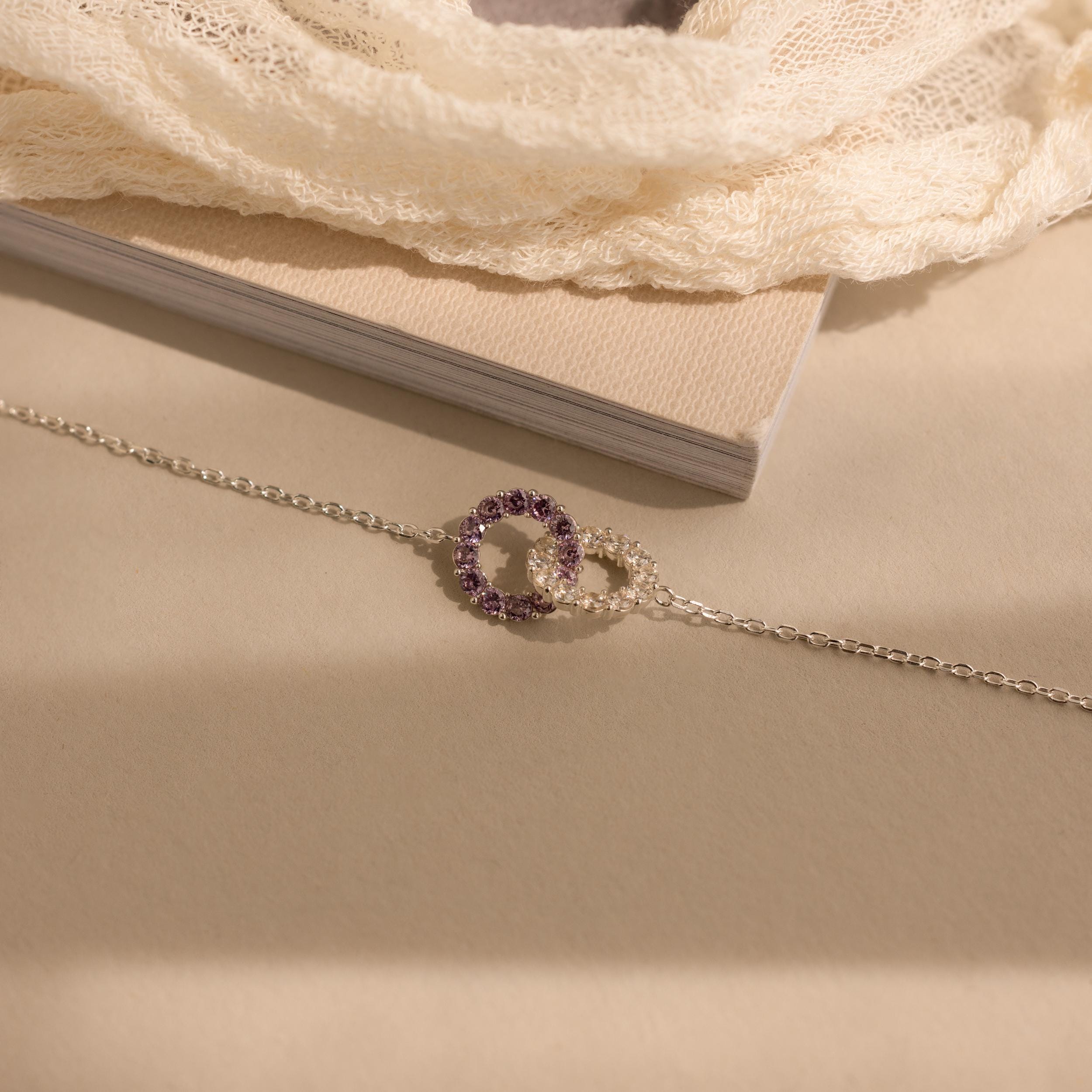 Interlocking Duo Birthstone Bracelet with two circles—one set with purple stones—displayed on a beige surface. This customizable bracelet is a meaningful gift for any occasion.