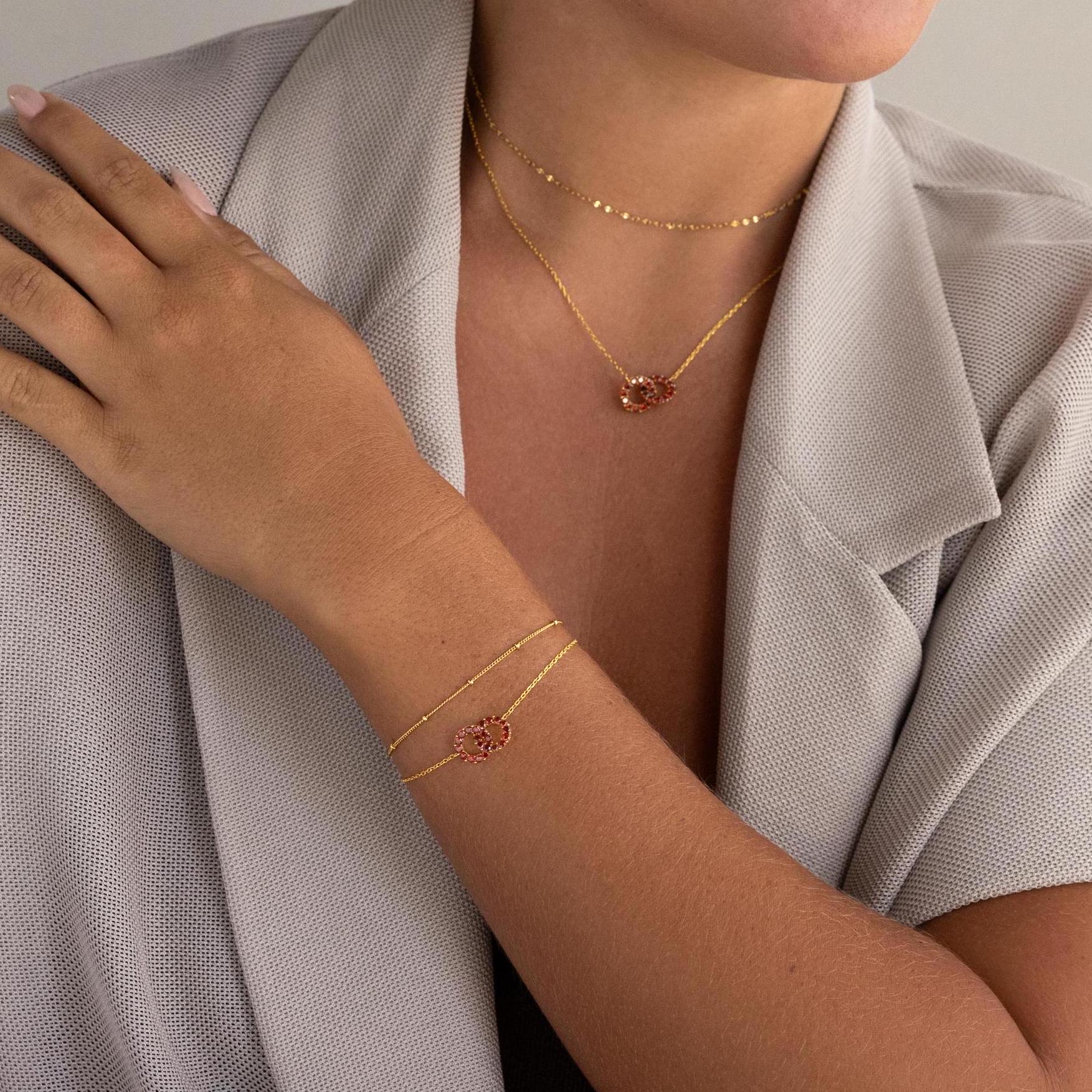 A woman in a light gray blazer and gold jewelry with pink flower charms rests her hand on her shoulder, highlighting the Interlocking Duo Birthstone Bracelet—a customizable gemstone piece ideal for meaningful gifting.