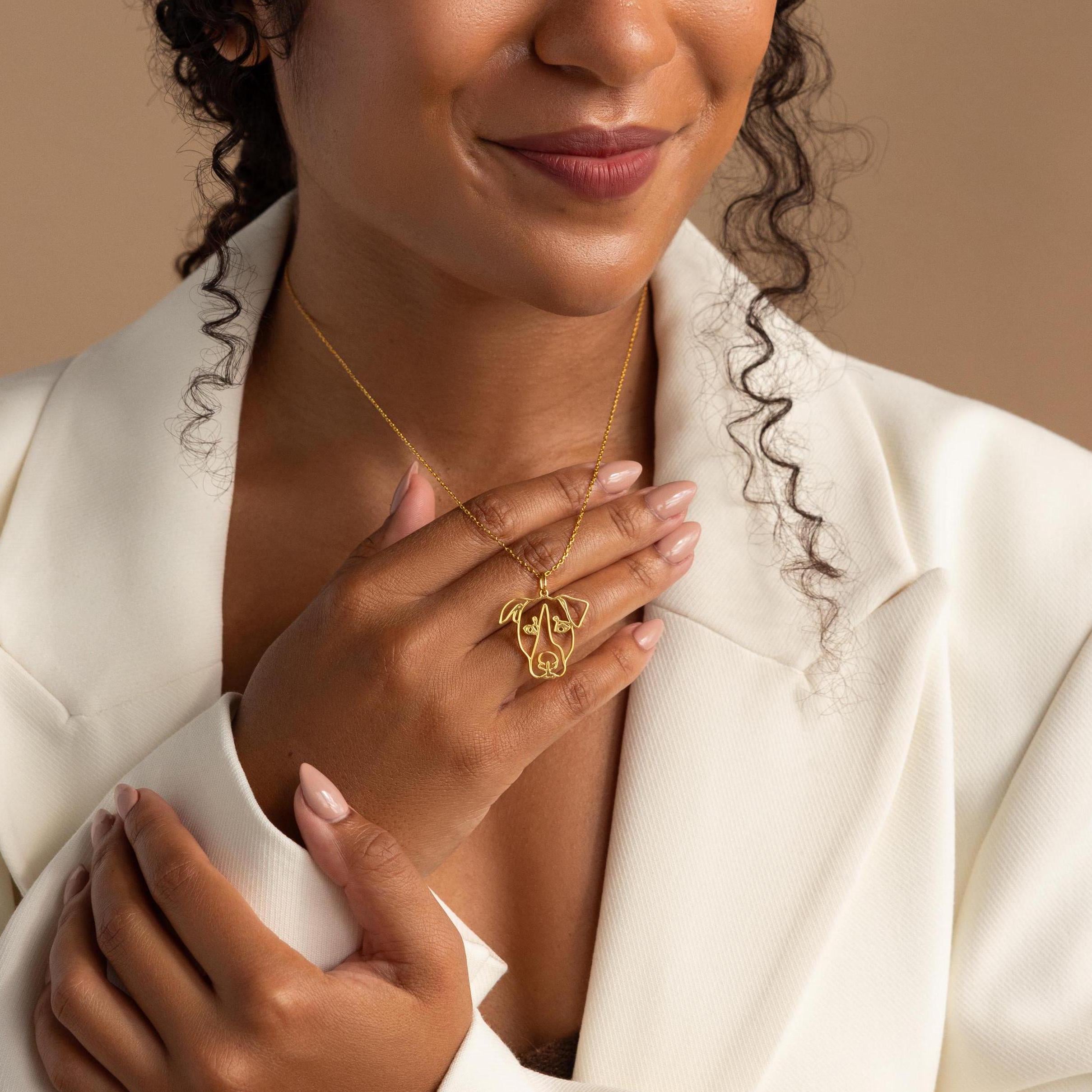 Woman in a white blazer holding a gold Custom Pet Line Art Necklace with an abstract face design, smiling softly.