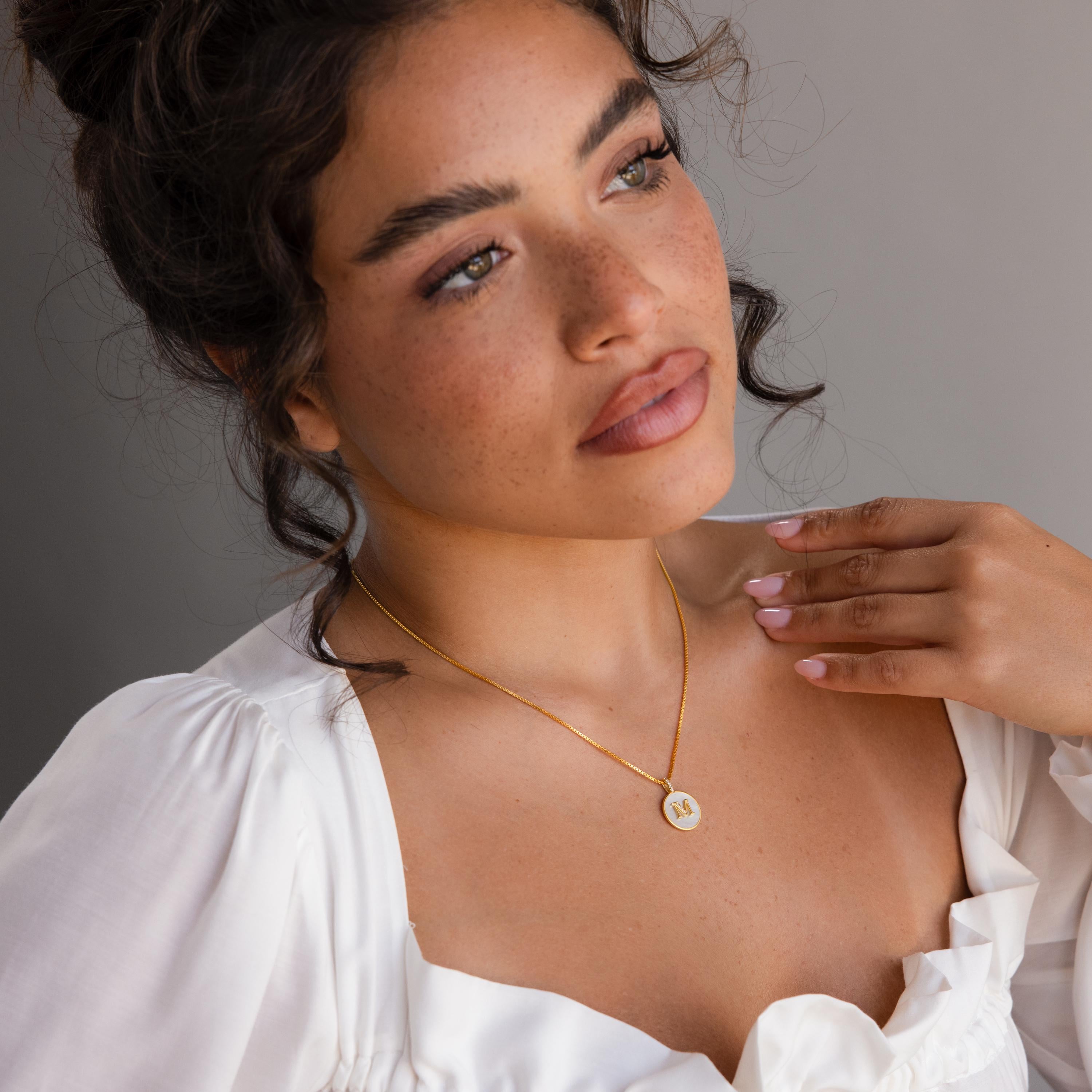 Woman with wavy dark hair in a white top, thoughtfully gazing to the side while wearing a Gemstone Initial Disc Necklace.