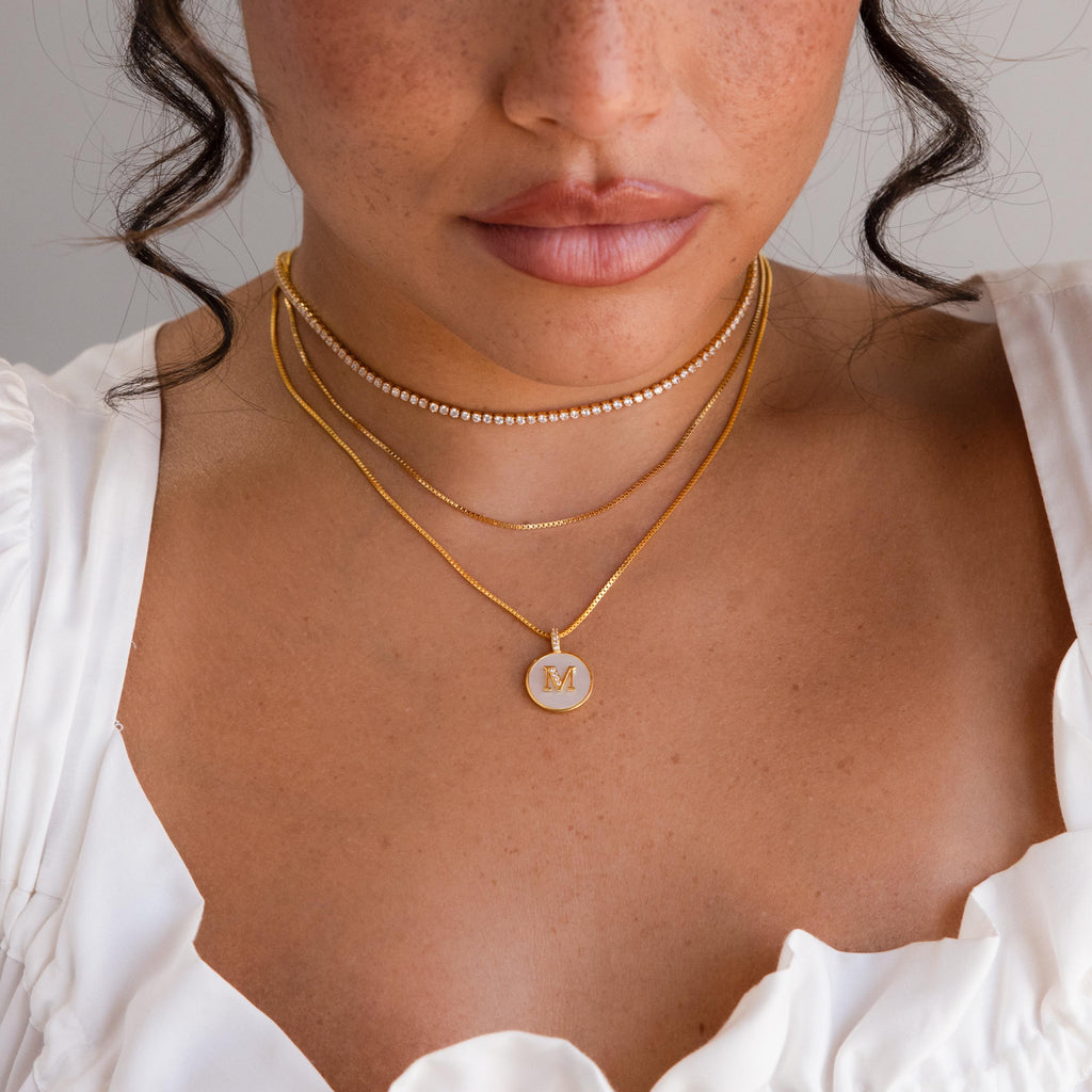 Woman wearing layered gold necklaces, including a Gemstone Initial Disc Necklace with an M pendant, paired with a white top featuring ruffled sleeves.