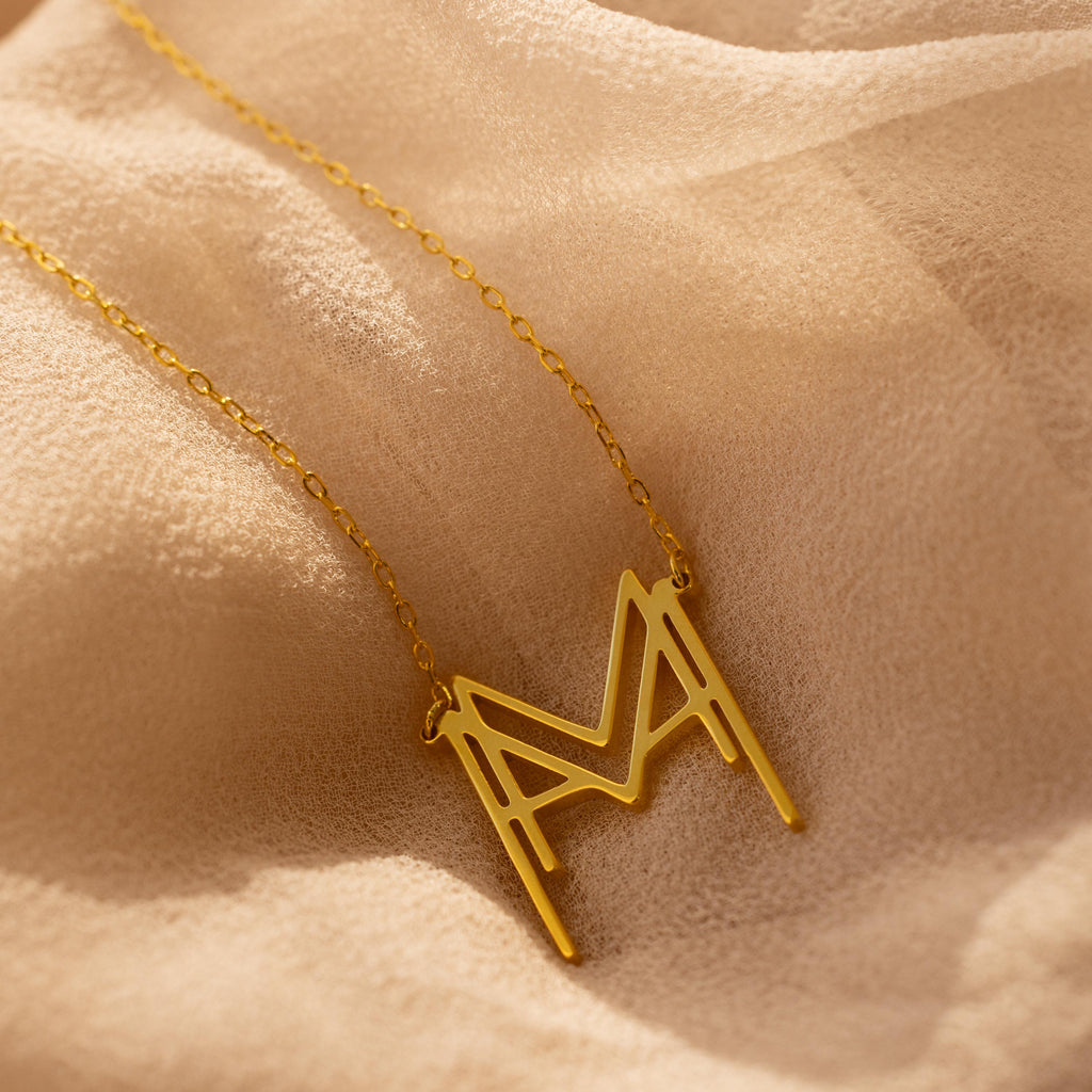The Modern Name Monogram Necklace with a large "M" pendant rests on soft beige fabric—ideal for fans of personalized, modern jewelry.