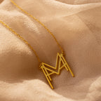 The Modern Name Monogram Necklace with a large "M" pendant rests on soft beige fabric—ideal for fans of personalized, modern jewelry.