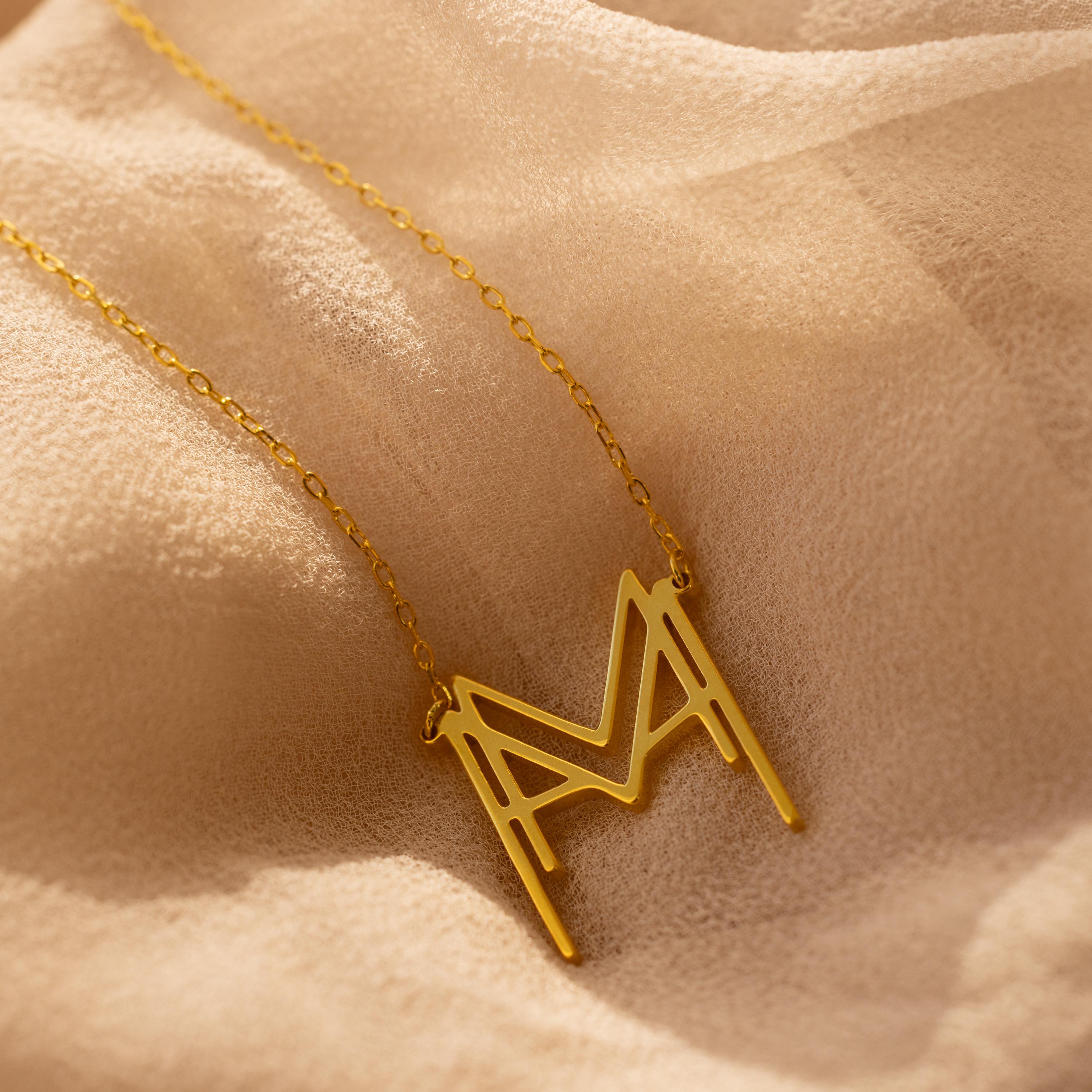 The Modern Name Monogram Necklace with a large "M" pendant rests on soft beige fabric—ideal for fans of personalized, modern jewelry.
