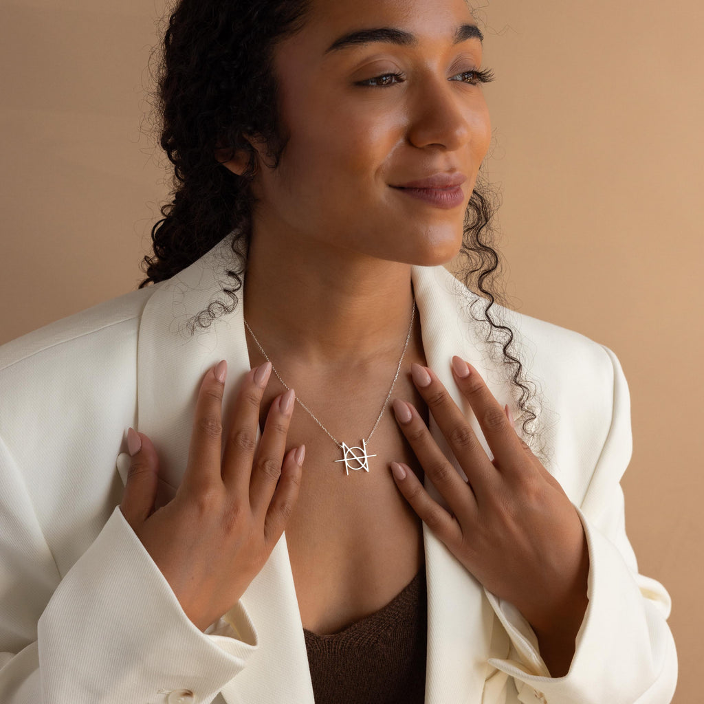 A smiling woman in a white blazer displays the Modern Name Monogram Necklace, featuring a personalized silver pendant, against a beige background.