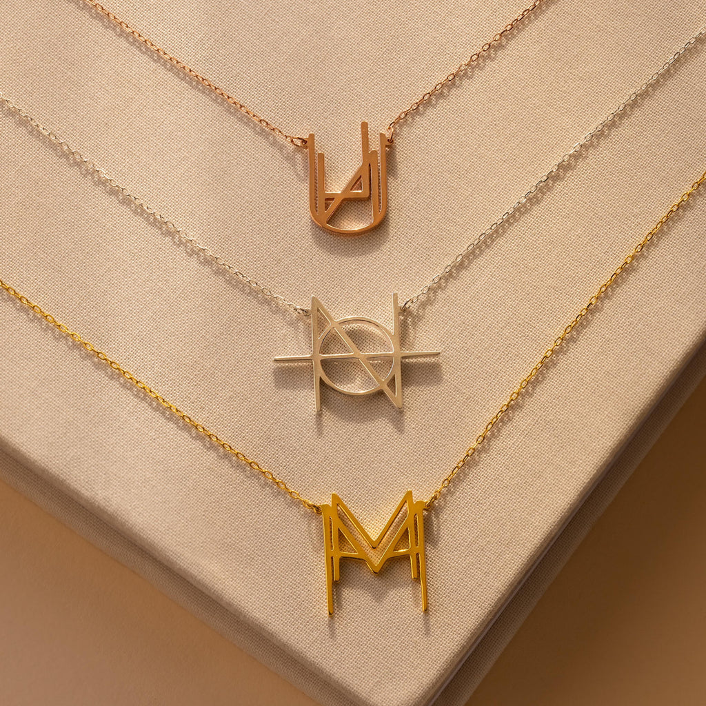 Three Modern Name Monogram Necklaces with unique metallic geometric pendants rest on a beige fabric background—ideal for those who love personalized jewelry and contemporary style.