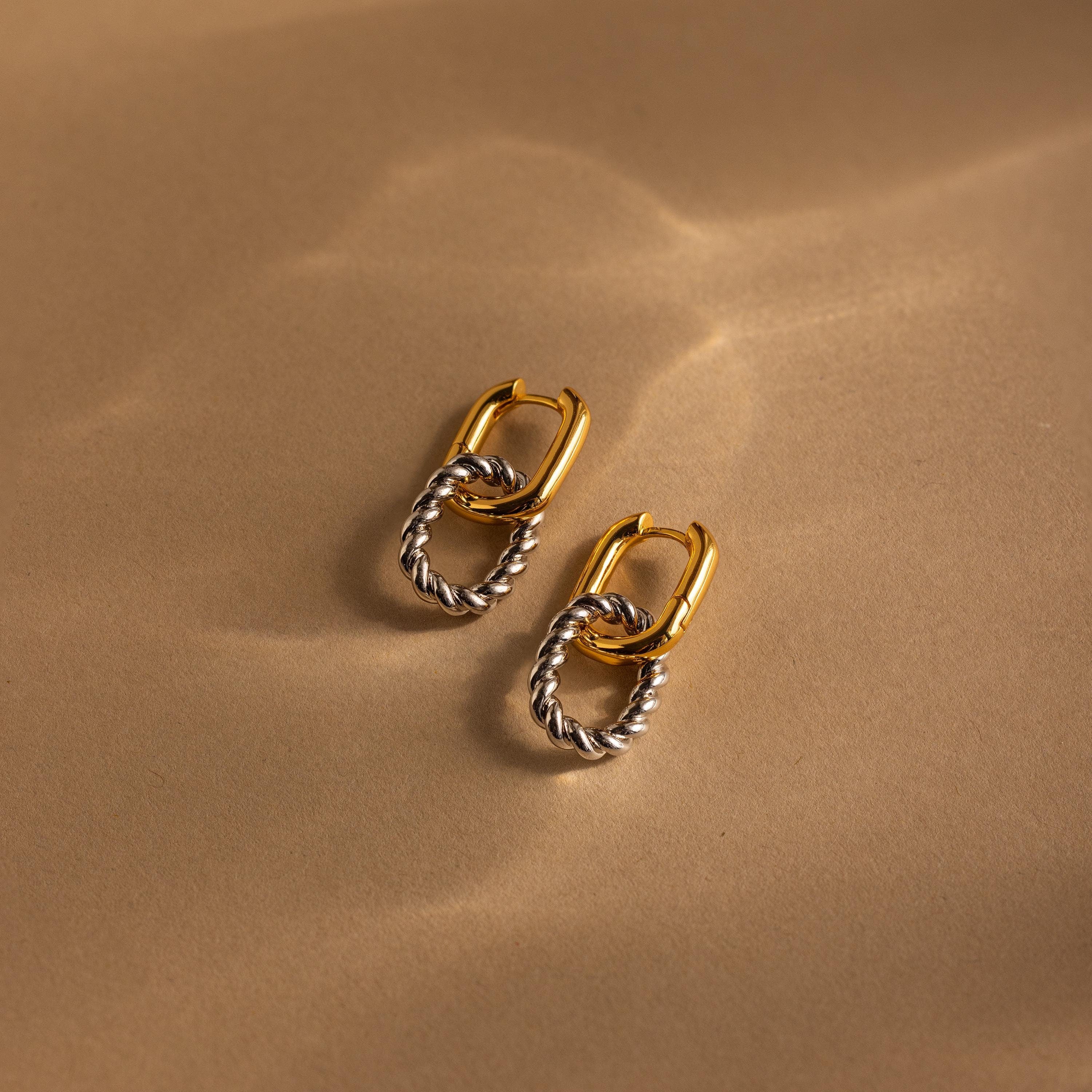 Mixed Metal Link Hoops displayed on a beige surface with soft light reflections, highlighting the elegant blend of gold and silver in these stylish earrings.