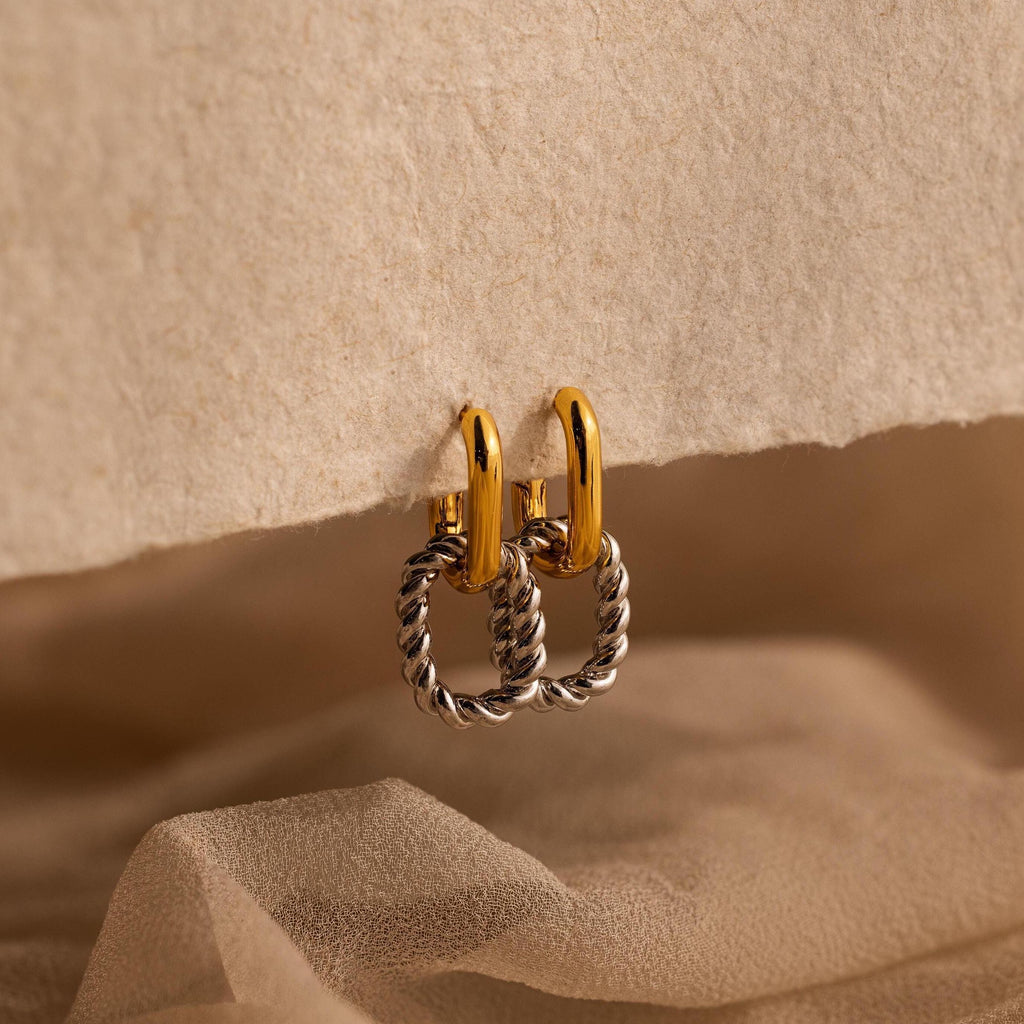 The Mixed Metal Link Hoops feature gold and silver in a twisted design, displayed on textured beige paper with a soft fabric background—chic earrings perfect for any style.