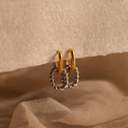 The Mixed Metal Link Hoops feature gold and silver in a twisted design, displayed on textured beige paper with a soft fabric background—chic earrings perfect for any style.