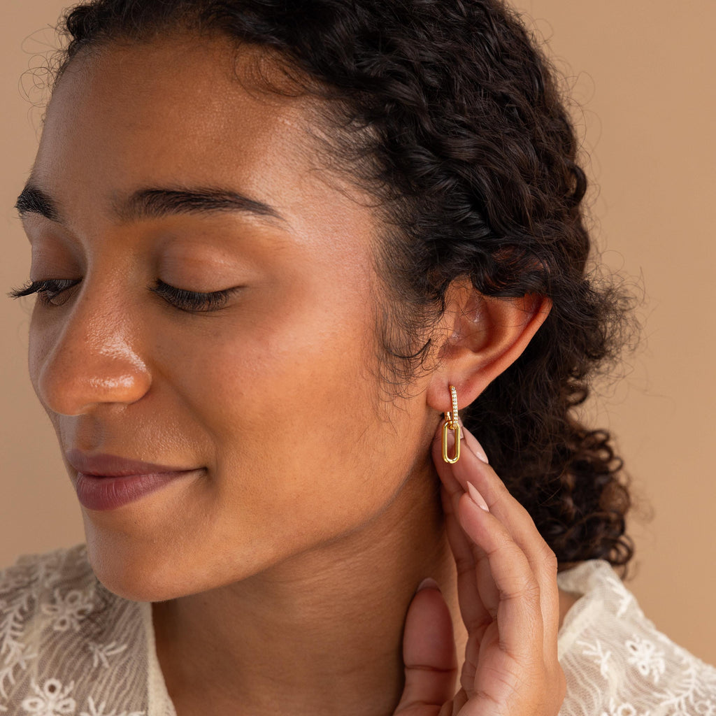 Woman with curly hair touches her Pave Link Drop Earrings, eyes closed, wearing a cream lace top against a beige background. Elegant CZ Diamonds accent these stylish earrings.