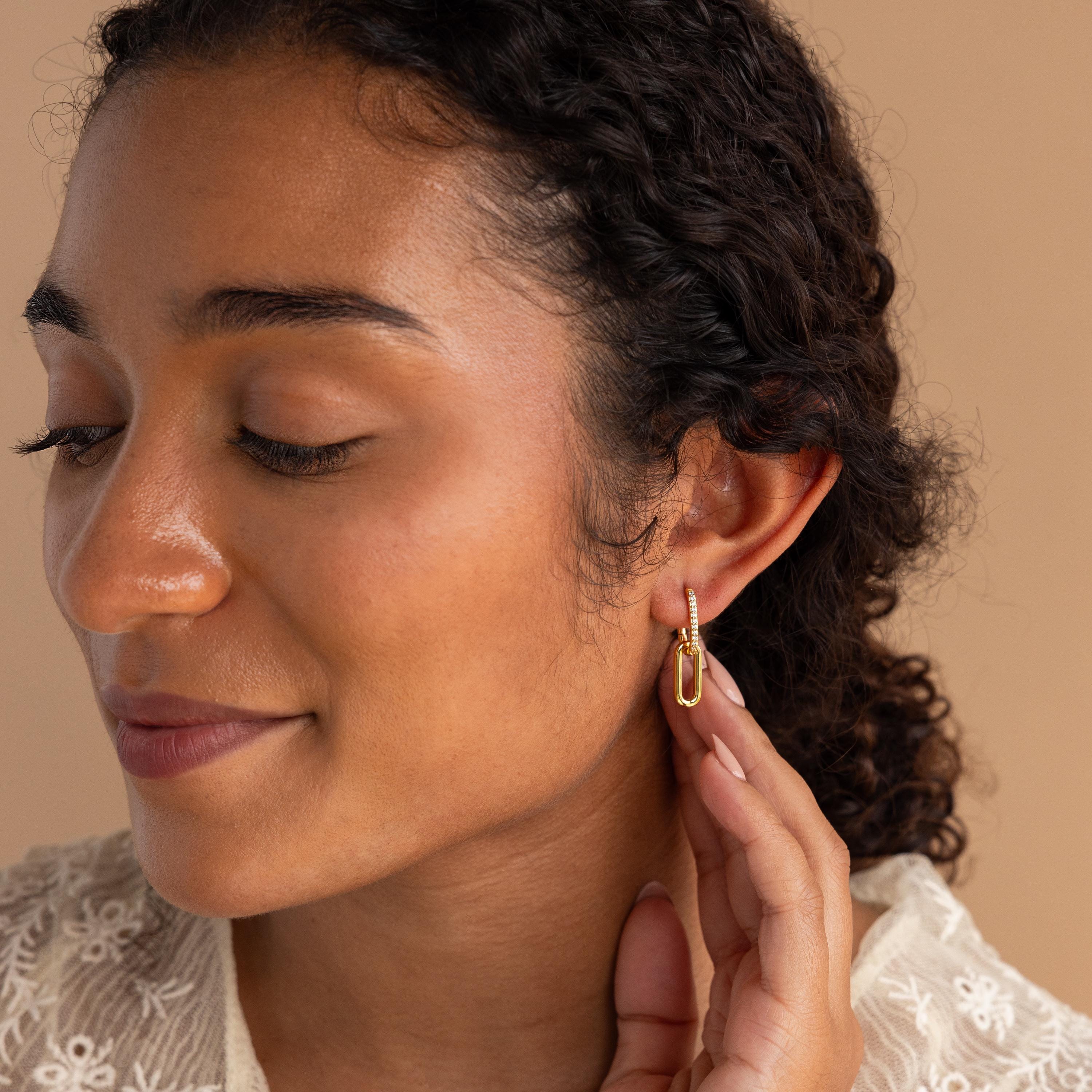 Woman with curly hair touches her Pave Link Drop Earrings, eyes closed, wearing a cream lace top against a beige background. Elegant CZ Diamonds accent these stylish earrings.