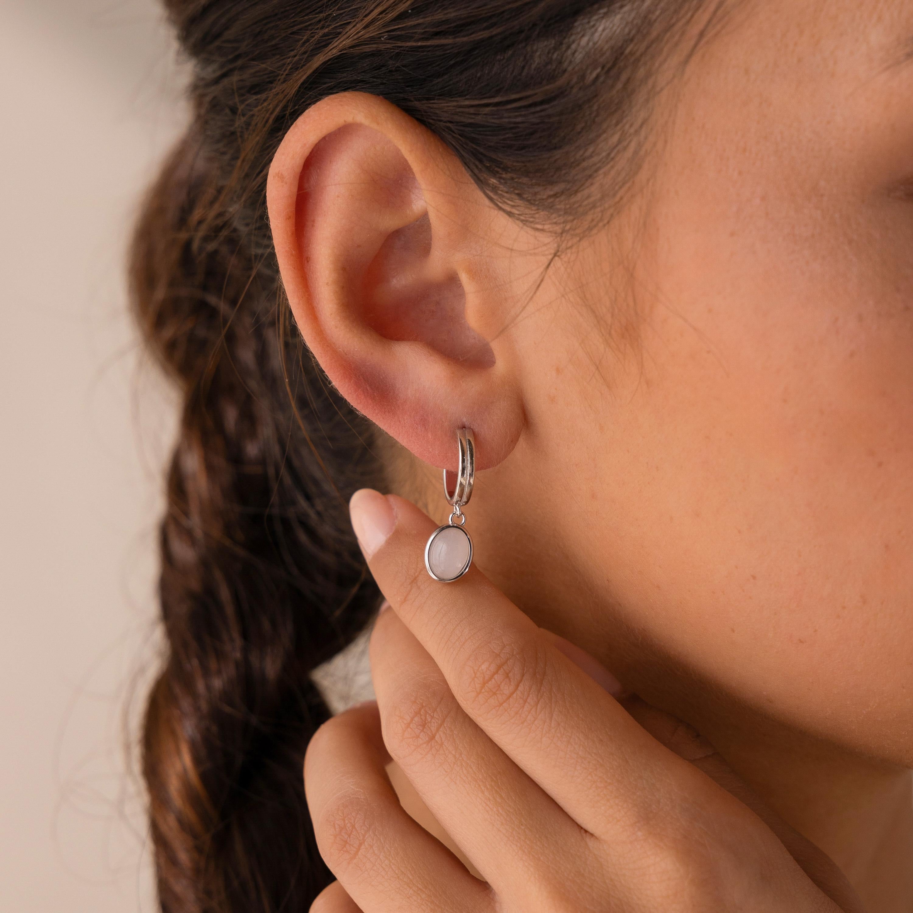A close-up shows a woman wearing the Moonstone Drop Hoops, her hand softly touching her earlobe to showcase these elegant silver hoops with white moonstone pendants.