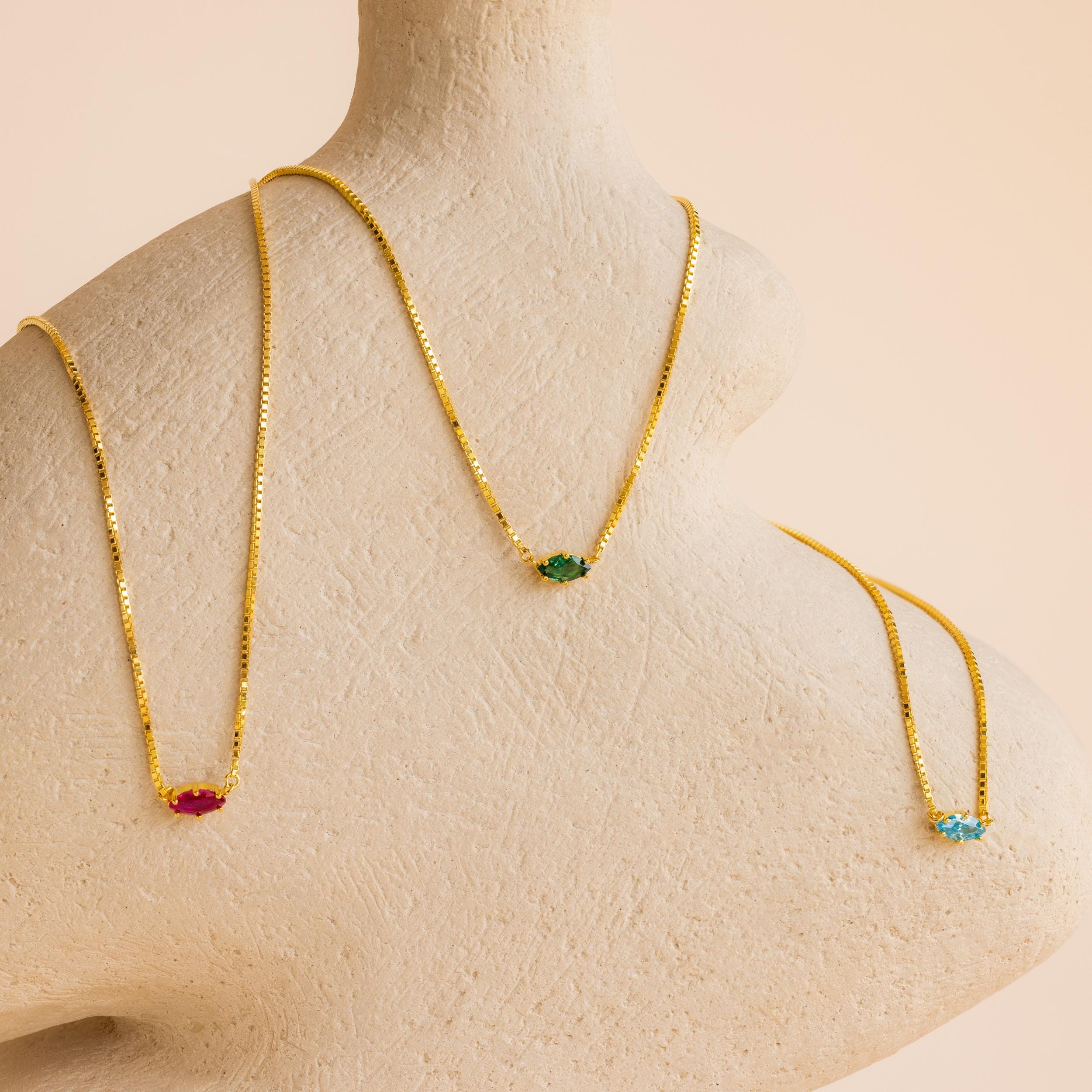 Three Dainty Marquise Birthstone Necklaces—red, green, and blue gemstone pendants—are shown on a beige bust with a light peach background. A perfect personalized jewelry gift for someone special.
