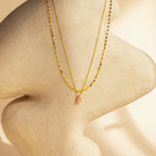 The Duo Layered Birthstone Necklace, a gold necklace featuring a delicate pendant, is displayed on a beige sculpted mannequin bust against a softly lit background.