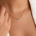 A person wears the Dainty Marquise Birthstone Necklace featuring a small blue marquise gemstone, shown close up against their bare neck and a white garment—a stunning piece of personalized jewelry.