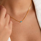 A person wears the Dainty Marquise Birthstone Necklace featuring a small blue marquise gemstone, shown close up against their bare neck and a white garment—a stunning piece of personalized jewelry.