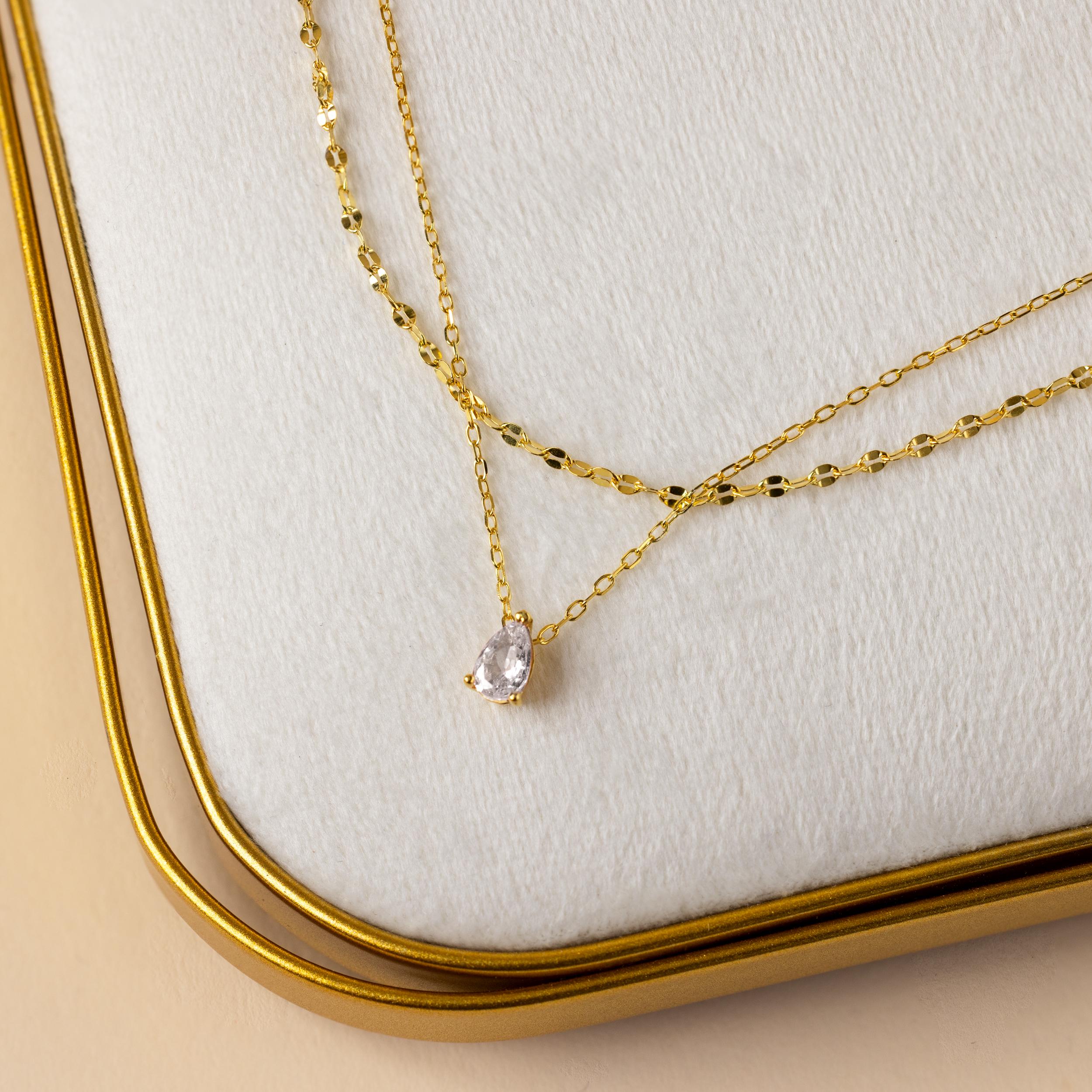 The Duo Layered Birthstone Necklace, featuring two delicate chains and a pear-cut birthstone, is displayed on an elegant gold-rimmed padded jewelry tray.