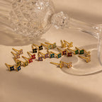 An Ivy Flower Birthstone Stud lies beside a crystal glass on its side, with gold and sterling silver stud earrings elegantly arranged on a beige surface.