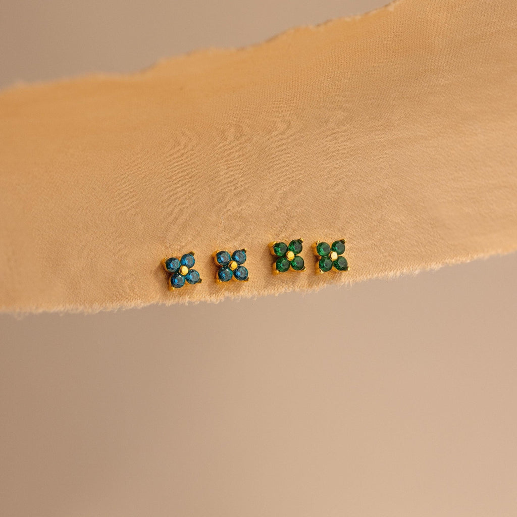 Four pairs of small stud earrings, including Ivy Flower Birthstone Studs featuring blue and green stones, are arranged in a row on a beige textured surface.