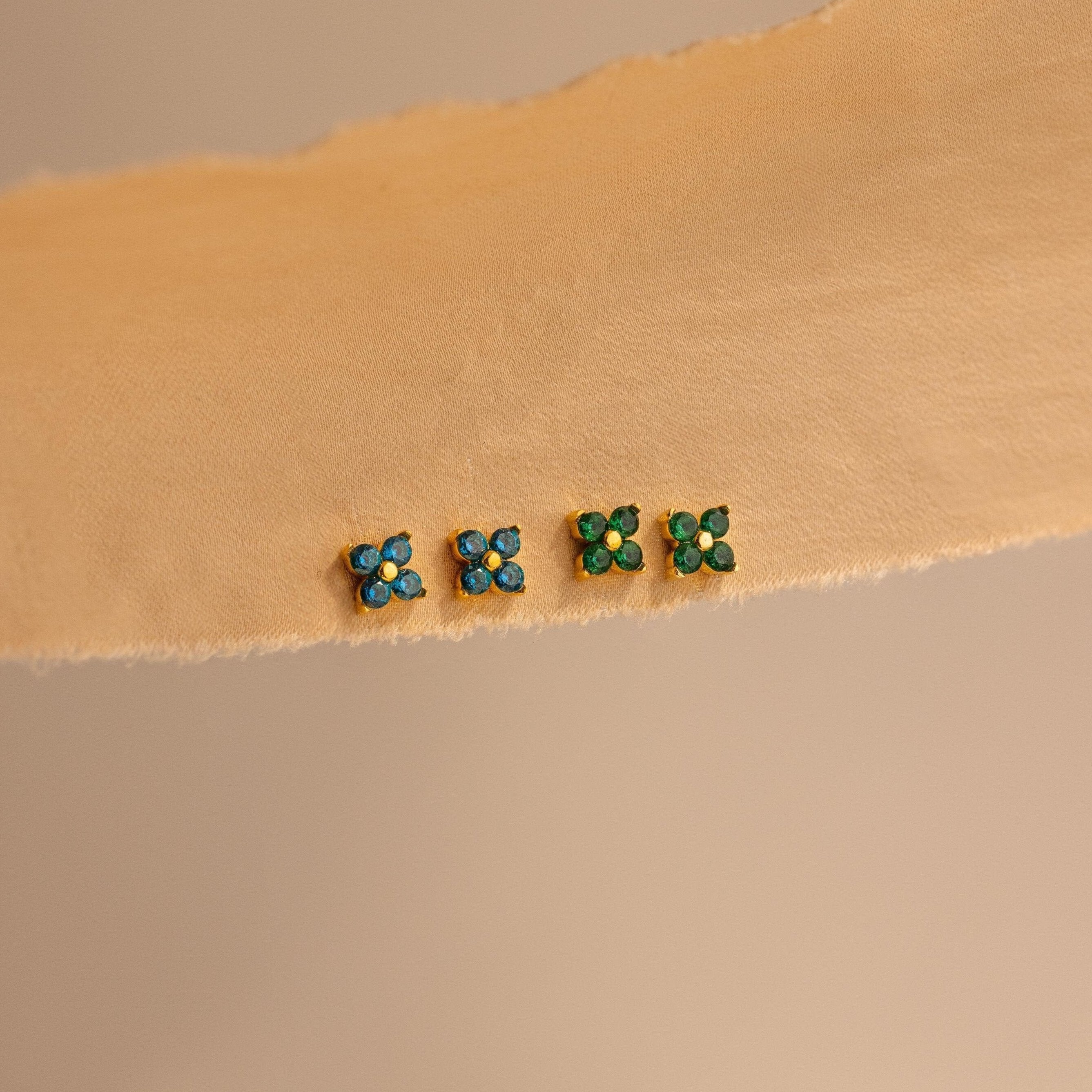 Four pairs of small stud earrings, including Ivy Flower Birthstone Studs featuring blue and green stones, are arranged in a row on a beige textured surface.