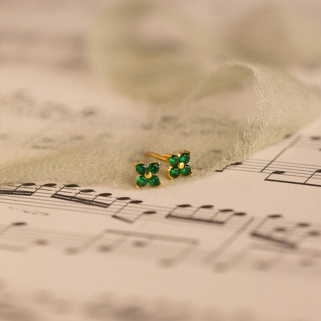 Delicate Ivy Flower Birthstone Studs, small green gemstone earrings, rest on sheet music with sheer fabric gently draped around them.