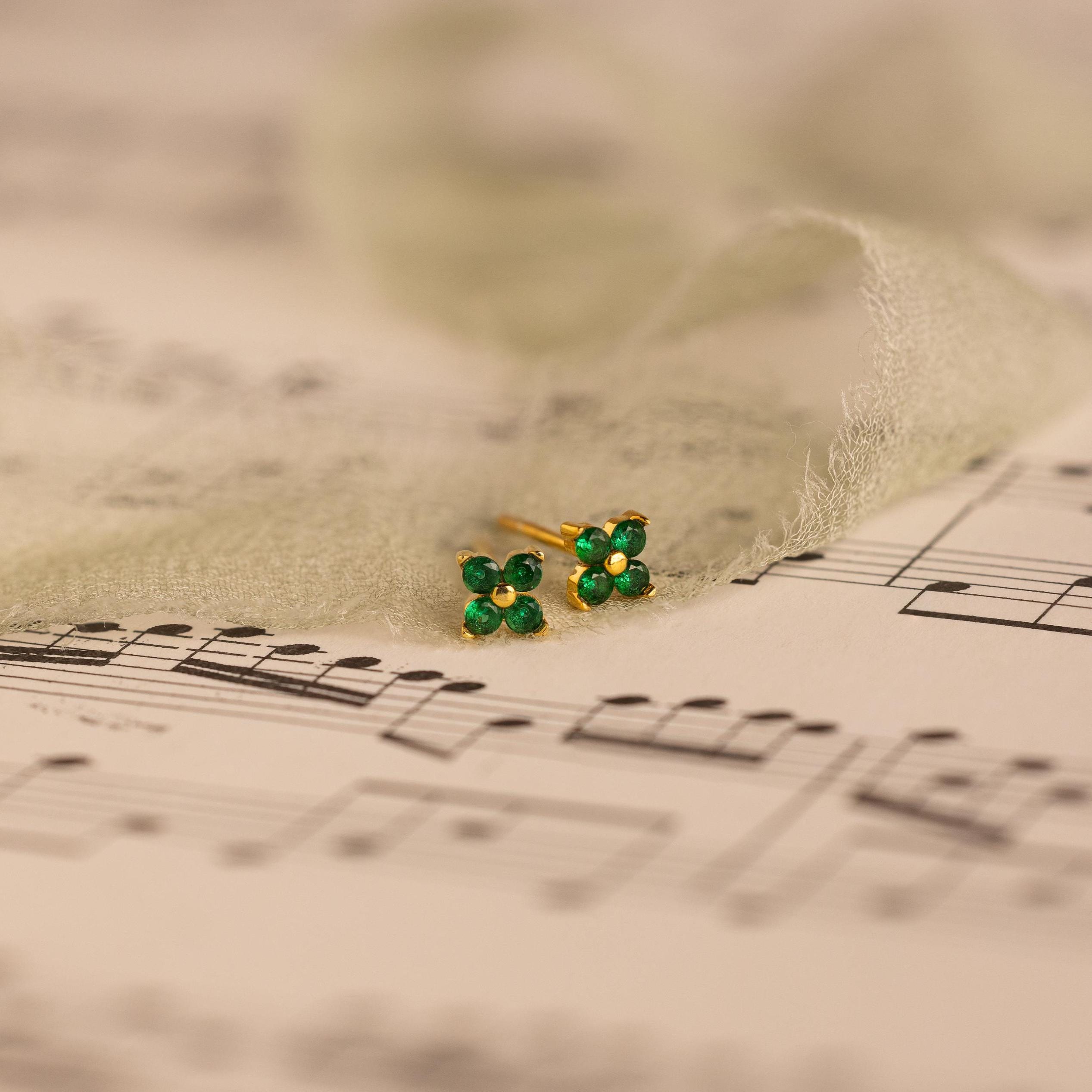 Delicate Ivy Flower Birthstone Studs, small green gemstone earrings, rest on sheet music with sheer fabric gently draped around them.