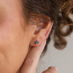 A close-up of a persons ear adorned with Ivy Flower Birthstone Studs in sterling silver, featuring a small blue gemstone, while a finger gently touches the ear.