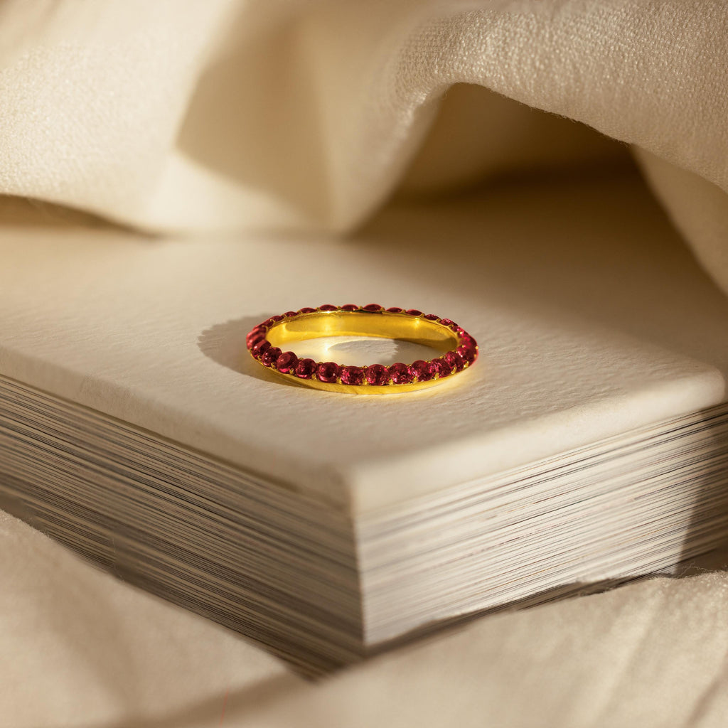 The Engraved Birthstone Eternity Ring, personalized and set with red gemstones, is displayed atop a closed book and surrounded by soft, cream-colored fabric.