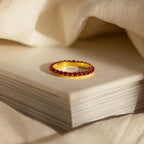 The Engraved Birthstone Eternity Ring, personalized and set with red gemstones, is displayed atop a closed book and surrounded by soft, cream-colored fabric.