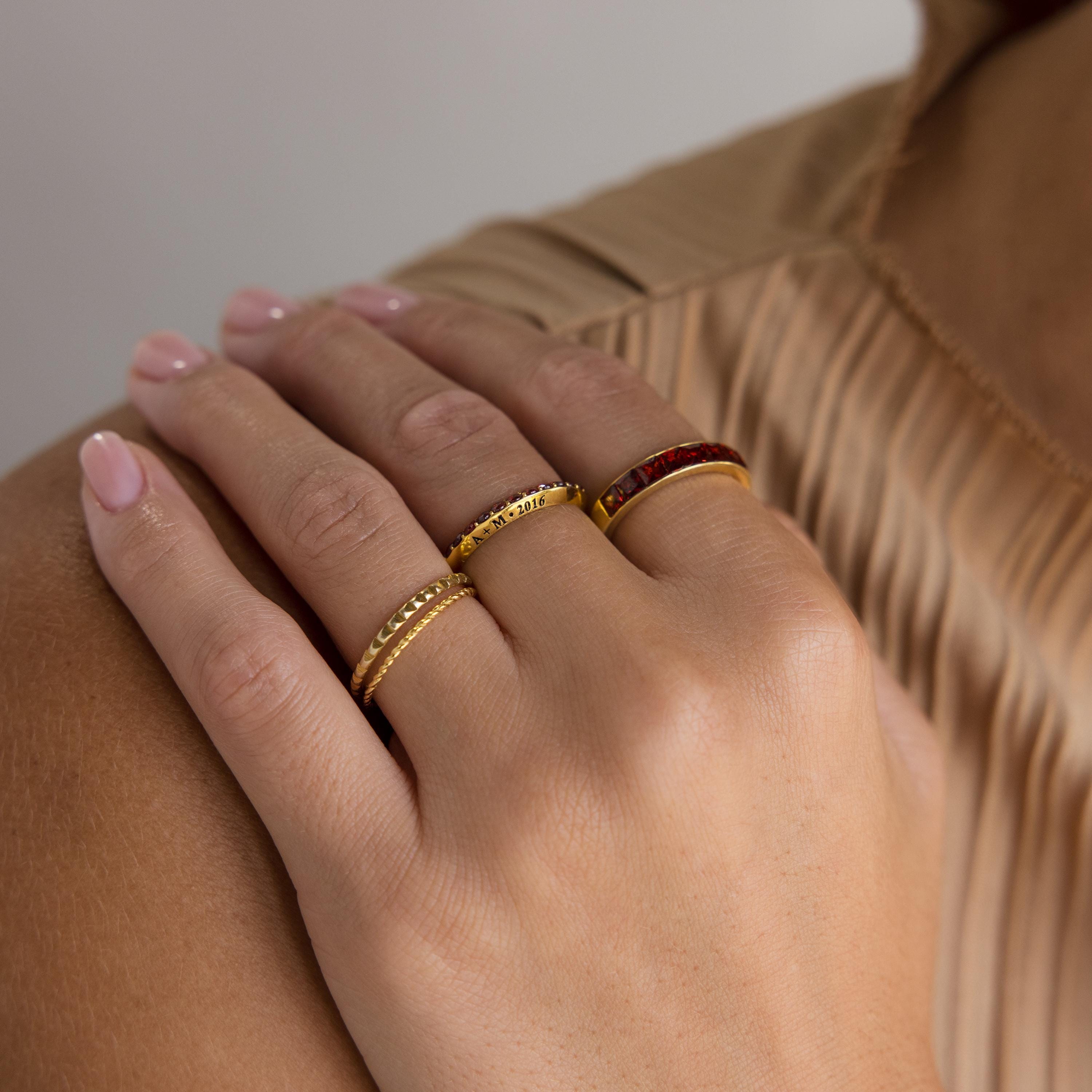 A hand with neatly manicured nails wears the Engraved Birthstone Eternity Ring and two gold bands, resting gracefully on beige fabric.