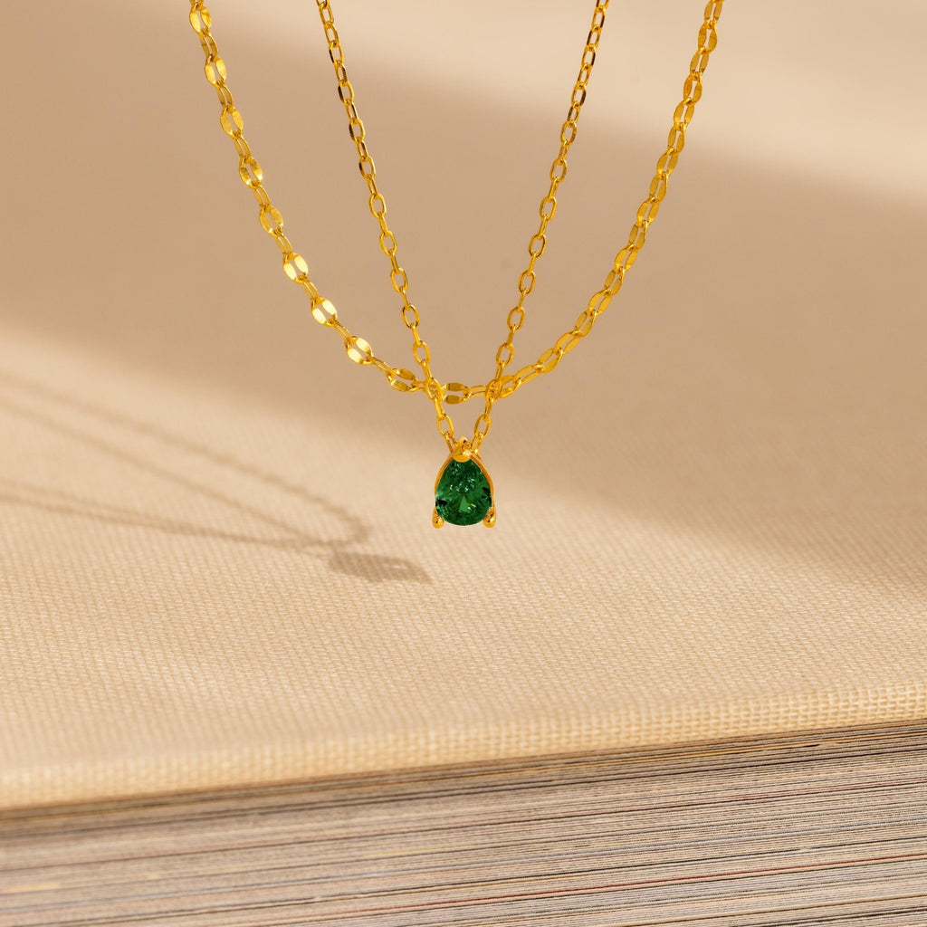 The Duo Layered Birthstone Necklace, showcasing a pear-cut green gemstone pendant, is displayed elegantly on a beige textured surface.
