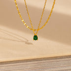 The Duo Layered Birthstone Necklace, showcasing a pear-cut green gemstone pendant, is displayed elegantly on a beige textured surface.