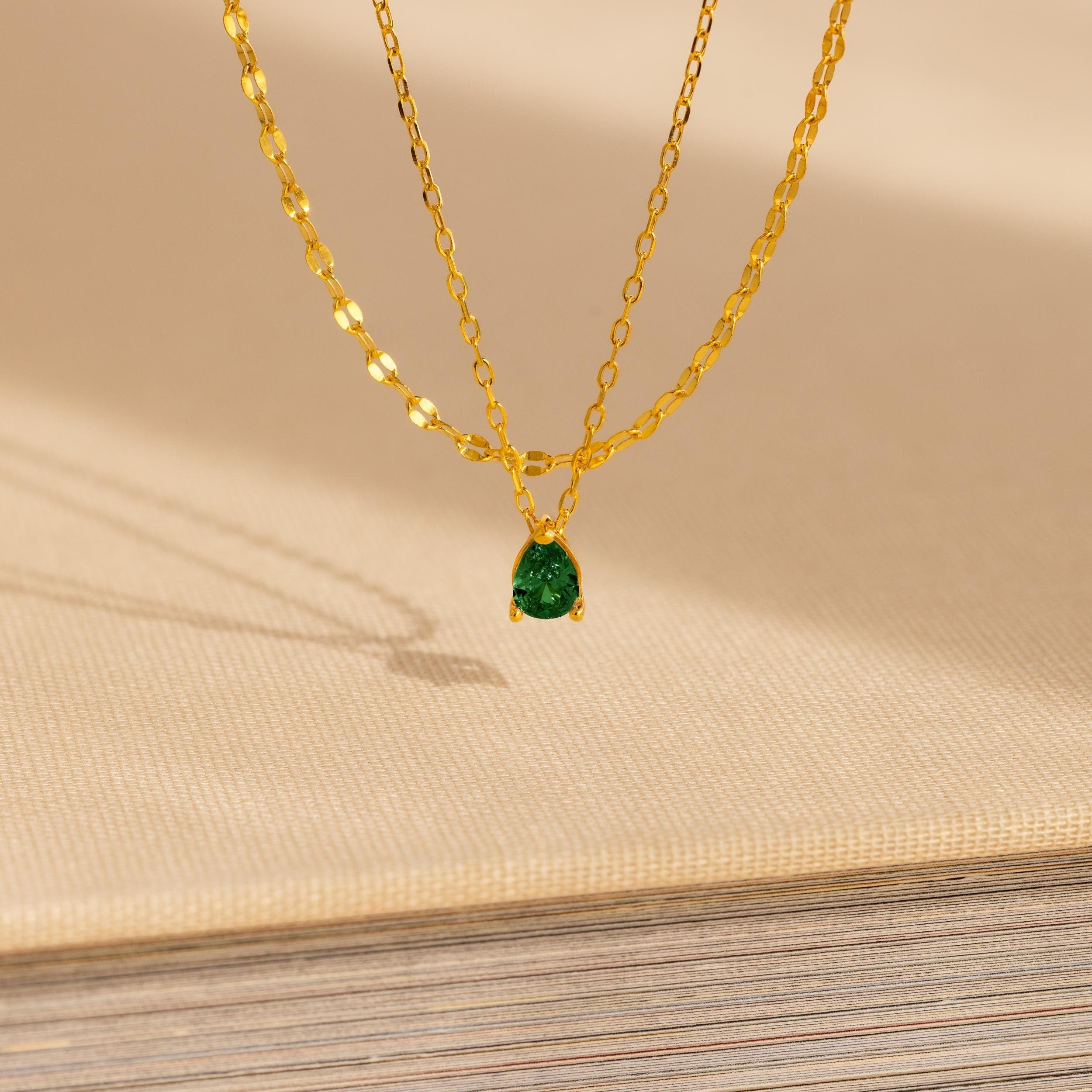 The Duo Layered Birthstone Necklace, showcasing a pear-cut green gemstone pendant, is displayed elegantly on a beige textured surface.