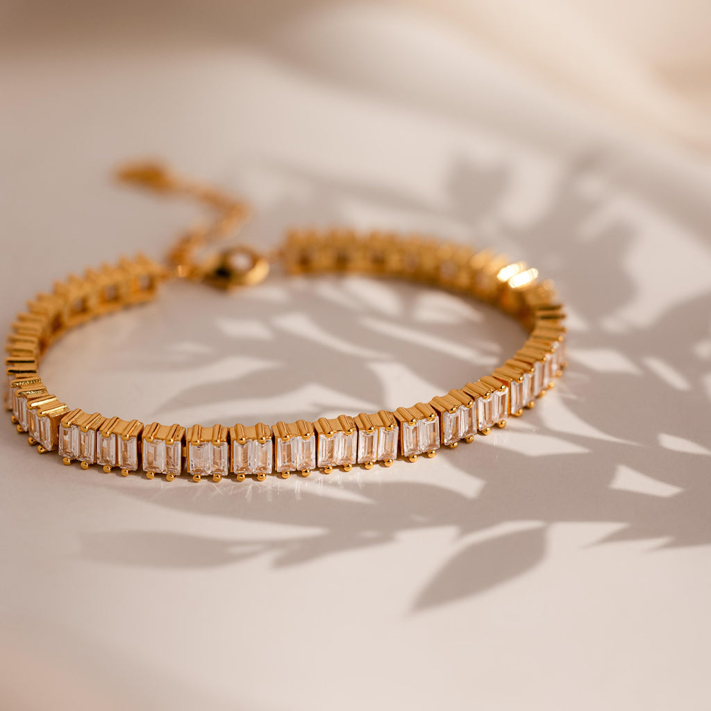 The Vertical Baguette Tennis Bracelet, featuring gold and rectangular gemstones evocative of baguette diamonds, casts a striking shadow on a light surface.