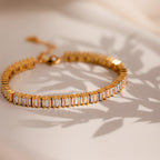 The Vertical Baguette Tennis Bracelet, featuring gold and rectangular gemstones evocative of baguette diamonds, casts a striking shadow on a light surface.