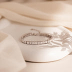 The Vertical Baguette Tennis Bracelet in silver rests on a white surface with soft beige fabric behind it, highlighting its elegant baguette diamonds and refined Art Deco-inspired charm.