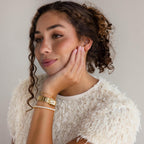 A woman with curly hair in a textured white top, resting her chin on her hand, wears the Vertical Baguette Tennis Bracelet in gold, adorned with sparkling baguette diamonds.