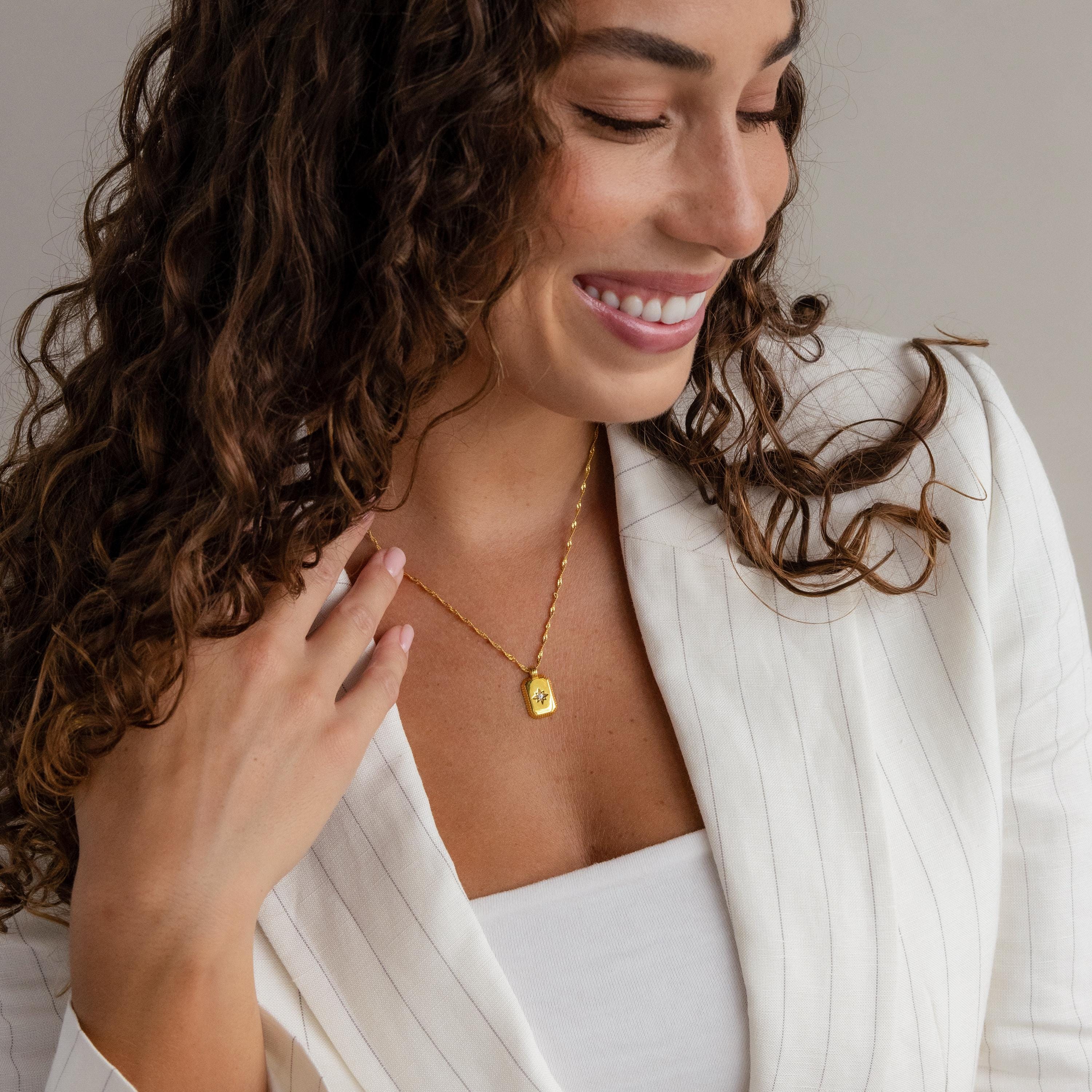 A woman in a white blazer smiles, wearing the Ribbed Birthstone Star Necklace and a gold pendant, gently touching her curly hair.