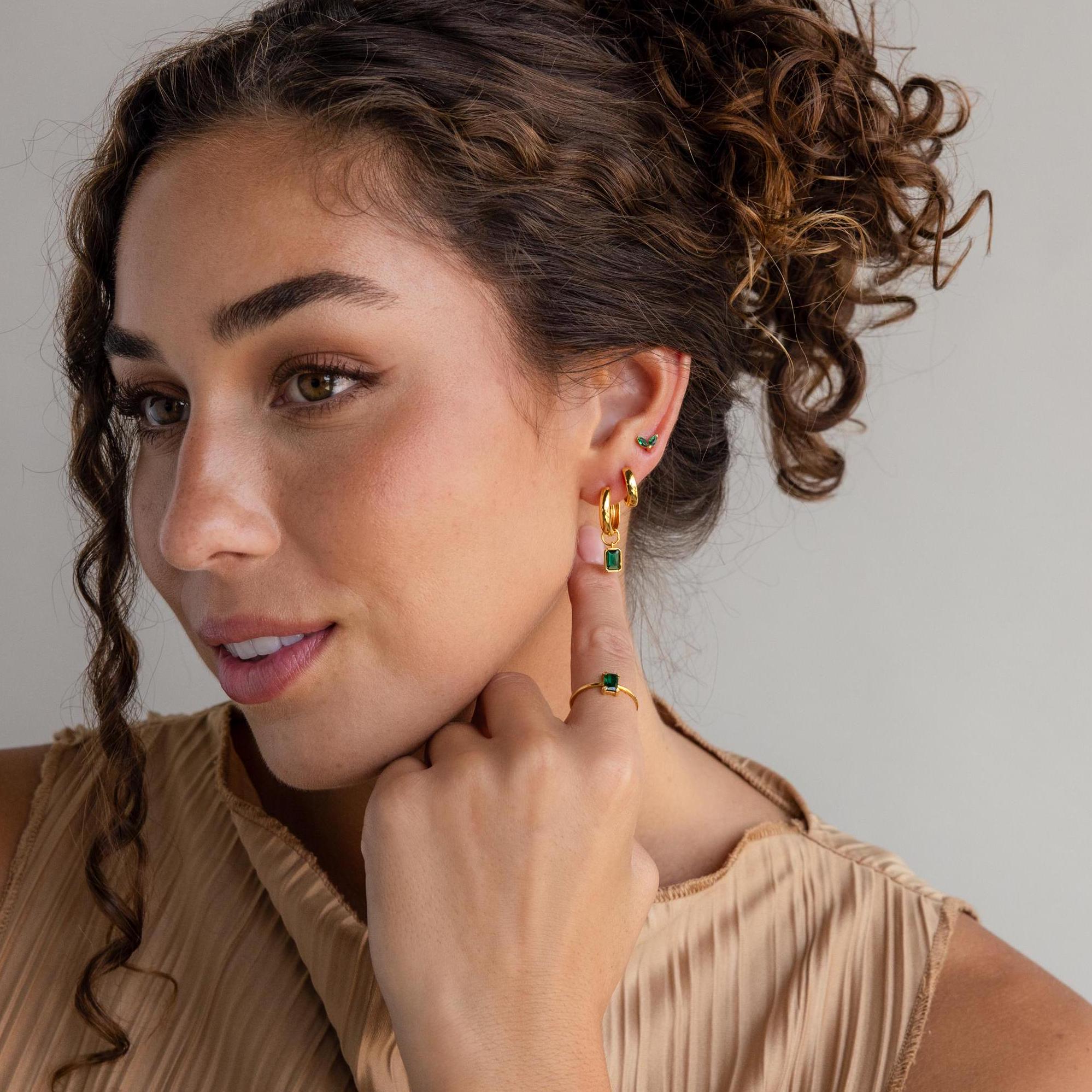 A woman with curly brown hair in a beige top and gold jewelry smiles, pointing to her Marquise Petal Birthstone Studs in Emerald (May), highlighting her love for personalized jewelry.