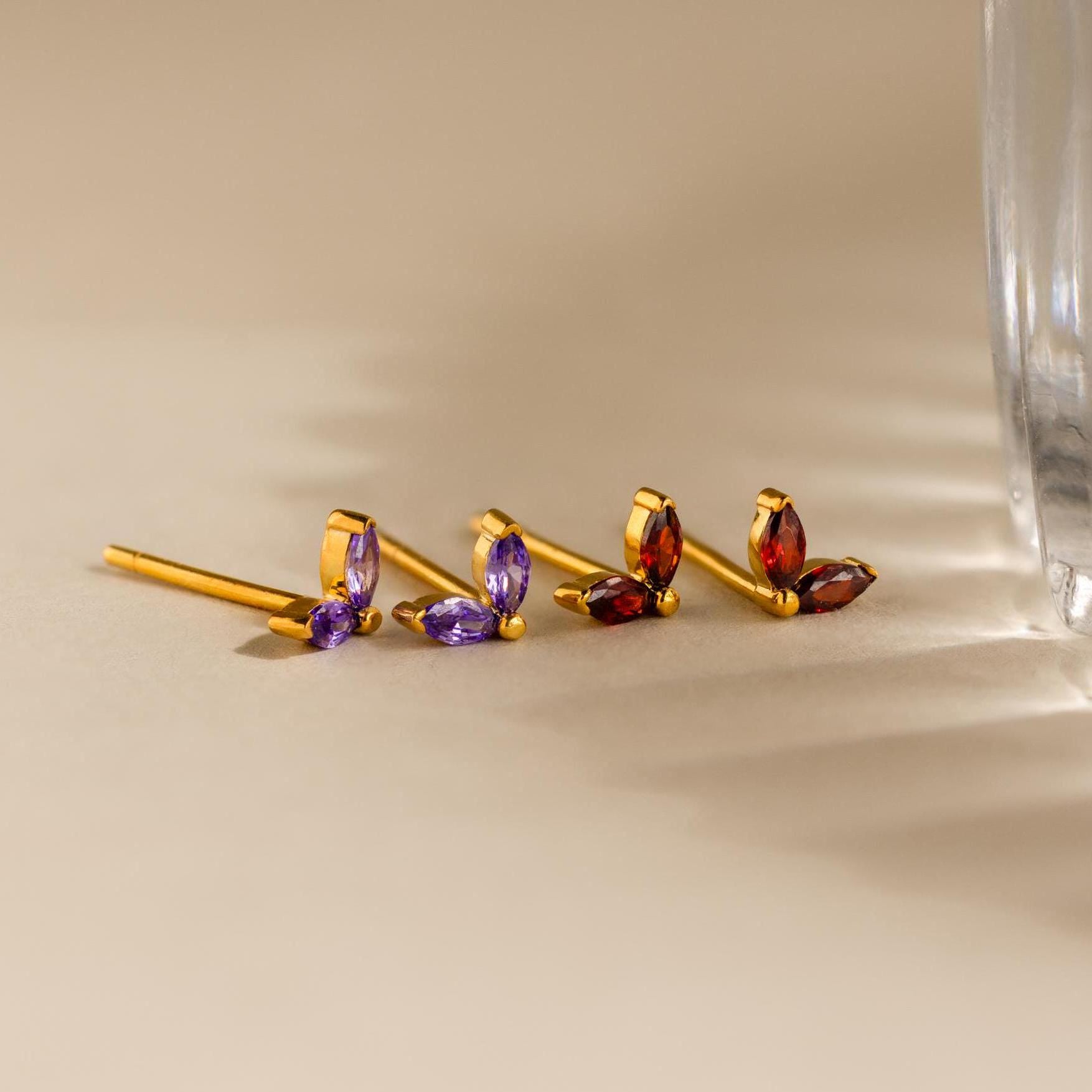 Marquise Petal Birthstone Studs in gold showcase two marquise gemstones—one pair purple, one red—displayed on a beige surface. These elegant studs are perfect as personalized birthstone jewelry.