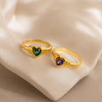 Two Birthstone Heart Signet Rings, featuring heart-shaped green and purple gemstones, rest on a soft beige fabric background.
