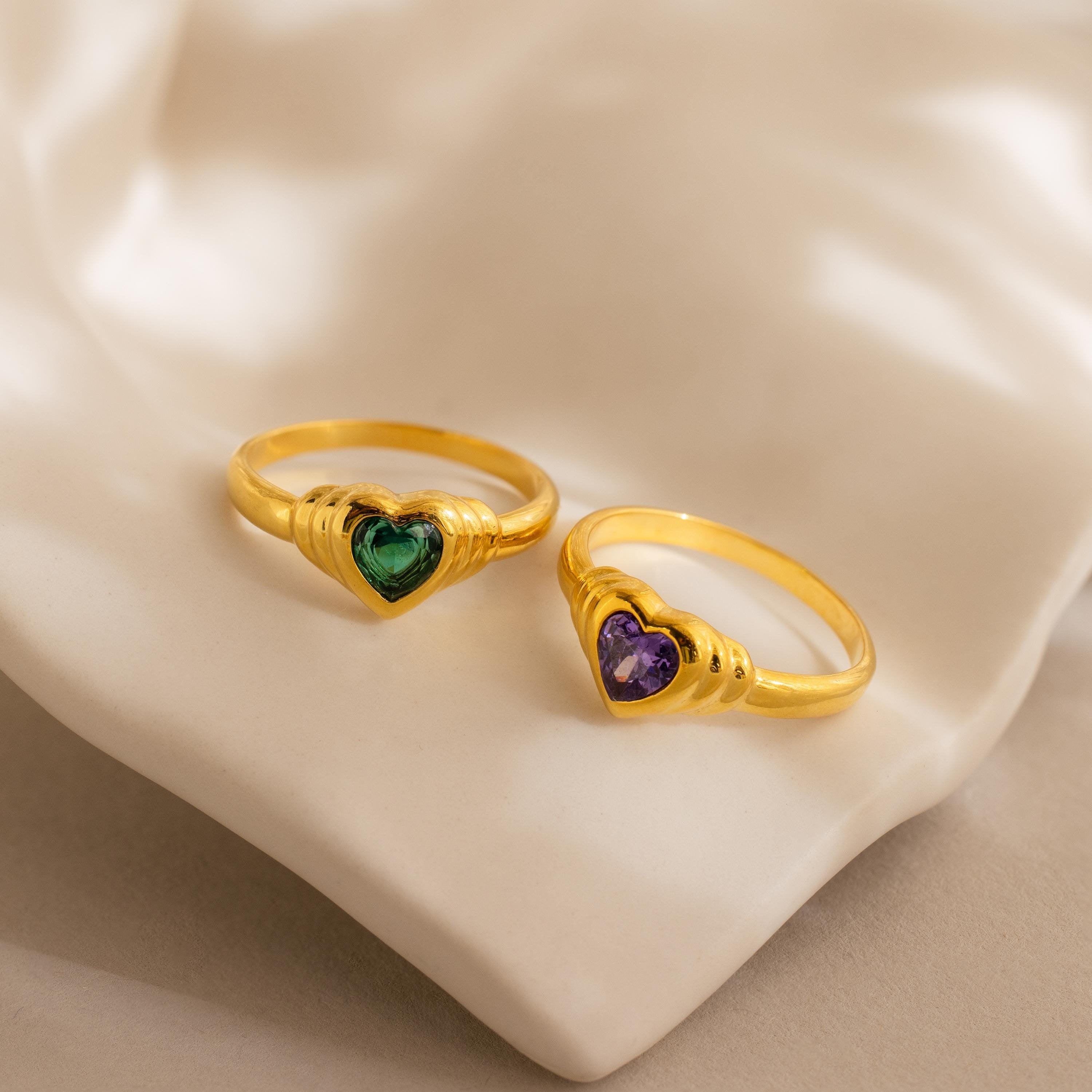 Two Birthstone Heart Signet Rings, featuring heart-shaped green and purple gemstones, rest on a soft beige fabric background.