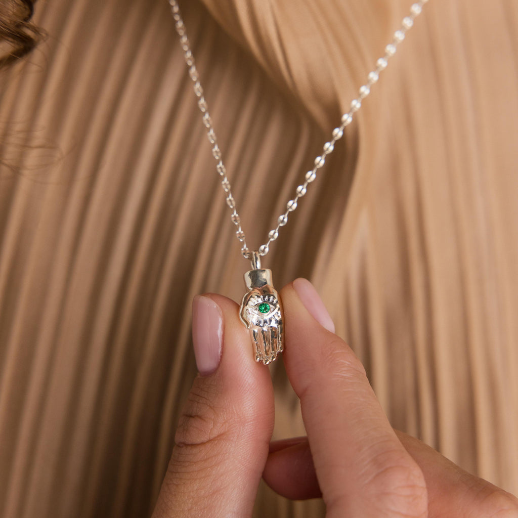 A hand holds the Evil Eye Birthstone Hand Necklace, showcasing a silver Hamsa pendant with a blue-green eye, blending protection and style.