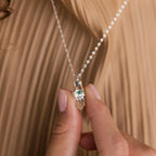 A hand holds the Evil Eye Birthstone Hand Necklace, showcasing a silver Hamsa pendant with a blue-green eye, blending protection and style.